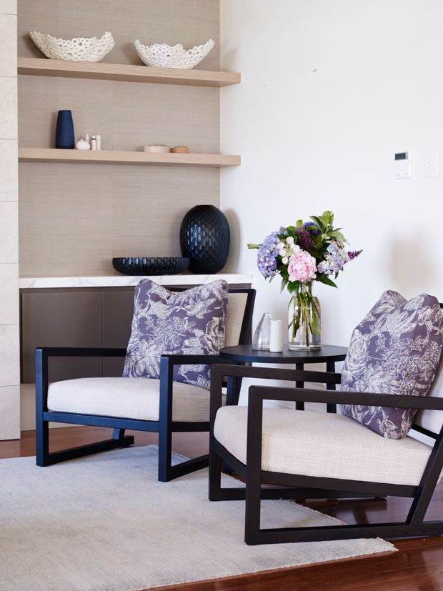 A refined sitting area with two dark timber-framed armchairs upholstered in light fabric and layered with patterned cushions, arranged around a small round black table. Built-in open shelving with a light timber or wood-look backing displays decorative ceramics and dark sculptural vases, while a large pale rug sits over dark timber flooring. A fresh floral arrangement adds colour to the otherwise neutral, contemporary interior.