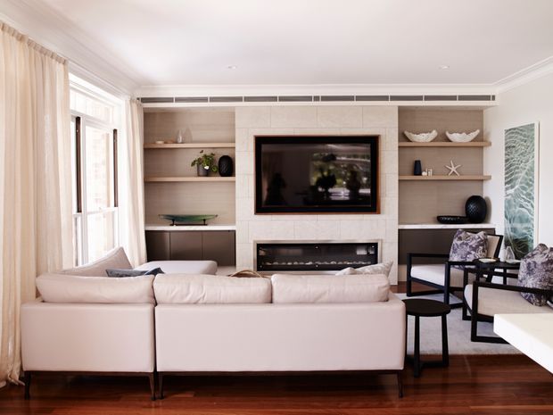 Elegant contemporary living room viewed from behind a large pale sectional sofa, with a built-in entertainment wall featuring a television, fireplace, open shelving, and integrated cabinetry. Tall windows with sheer curtains line the left side, while dark timber flooring, light upholstery, and black-framed occasional chairs create a refined, warm interior.