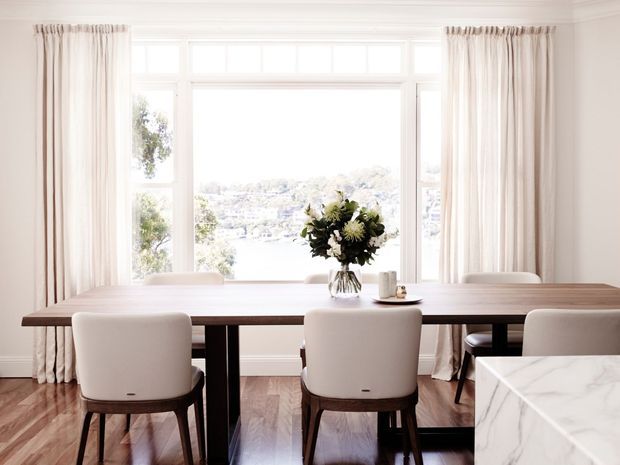 Bright, refined dining area with a long timber dining table and upholstered dining chairs positioned in front of large multi-pane windows. Full-height light curtains frame a scenic water view, while a marble-clad surface is visible in the foreground. The room has polished timber flooring, white walls and trim, and understated contemporary styling.