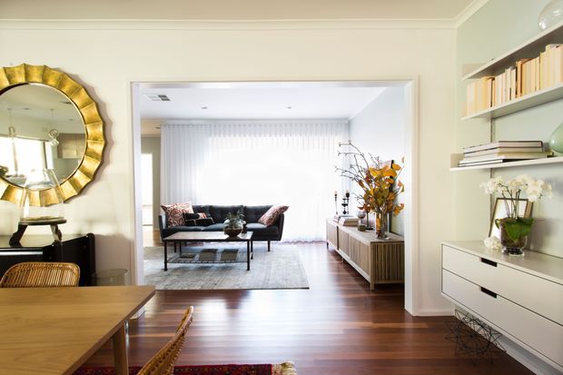 A bright, refined living and dining area with an open connection between the spaces. The foreground features a timber dining table and woven chairs, while the adjoining living room includes a dark sofa, coffee table, rug, and low timber media console. Built-in white shelving and drawers line the right wall, with a large gold-framed round mirror and decorative styling on the left. Warm timber flooring, pale walls, and sheer curtains create an elegant, welcoming interior.