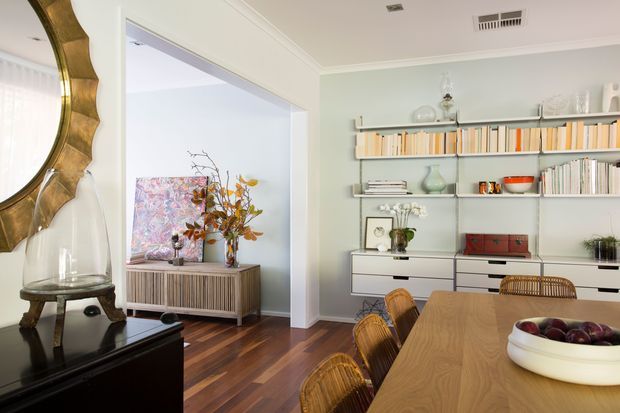 A refined open-plan dining and living area with a long timber dining table, woven rattan dining chairs, and a large white serving bowl. The room features pale green walls, dark timber flooring, white built-in cabinetry, and open shelving displaying books and decorative objects. A black sideboard and large sculptural gold-framed mirror sit to the left, with a bright adjoining sitting area visible through a wide opening.