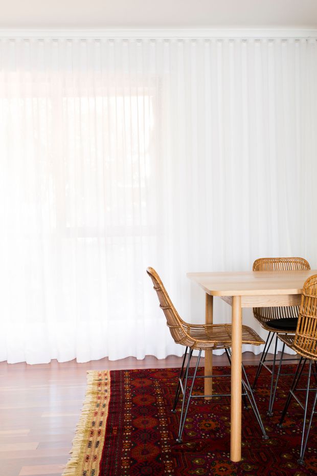 Bright dining area with full-height sheer white curtains, pale timber flooring, a simple light-wood dining table, and woven rattan dining chairs. A richly patterned red and burgundy Persian-style rug adds contrast beneath the dining setting.