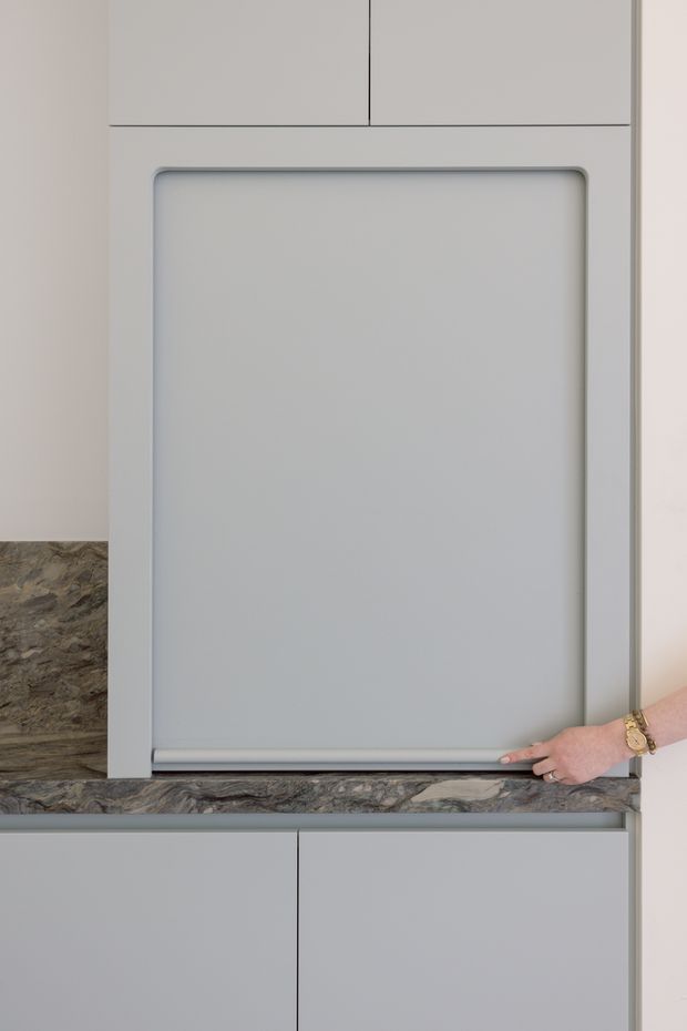 Close-up of a minimalist grey kitchen or utility cabinetry installation, featuring a large integrated roll-front panel or appliance door above a dark, heavily veined stone benchtop. Smooth light-grey cabinet fronts and clean recessed detailing create a contemporary built-in appearance; a hand is shown demonstrating the lower edge mechanism.