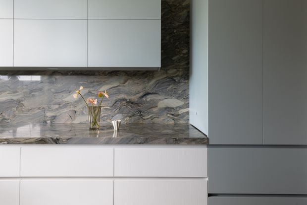 Close-up view of a refined contemporary kitchen with extensive grey veined natural-stone surfaces, including the benchtop and full-height splashback. Minimal white upper and lower cabinetry contrasts with tall dark grey integrated cabinetry on the right. A small vase provides a subtle decorative accent.