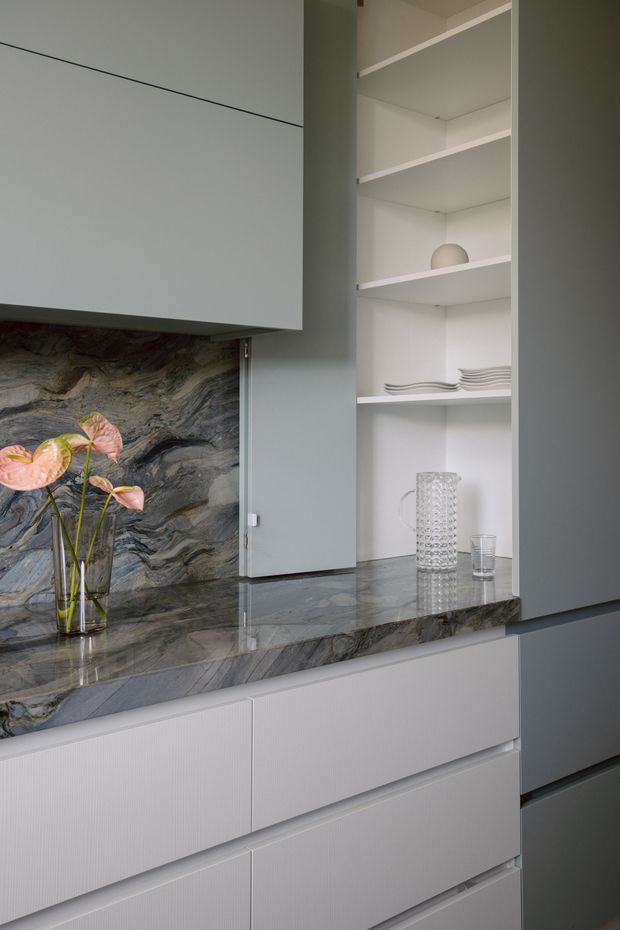 Close-up view of a refined kitchen or dining storage fit-out, featuring pale grey flat-front cabinetry, open white display shelving, and a dramatic dark veined natural-stone benchtop and splashback. Glassware, stacked plates, and a vase of flowers provide restrained styling accents.