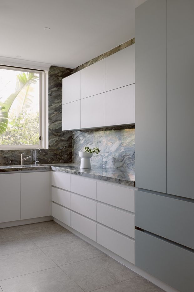 Bright contemporary kitchen with full-height matte grey cabinetry, white handleless wall and base cupboards, a dramatic veined stone backsplash and matching benchtop, integrated appliances, and large grey floor tiles. A window brings in abundant natural light and overlooks tropical foliage.