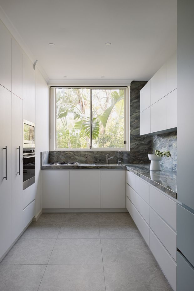 A compact, contemporary galley kitchen with white floor-to-ceiling cabinetry, integrated appliances, a dark grey veined stone benchtop and matching splashback, and a central window overlooking lush greenery. The room has a clean, minimal composition with large-format grey floor tiles, recessed ceiling lights, and a peninsula-style work surface.