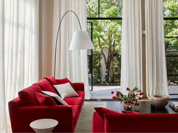 A bright, refined living room with vivid red sofas, white cushions, sheer floor-to-ceiling curtains, and a tall arched floor lamp. Large black-framed glazing opens views to a leafy courtyard, while a low dark coffee table displays a floral arrangement and decorative objects. The space has a strong red-and-white contrast and abundant natural light.