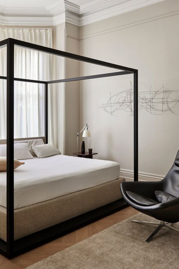 A refined bedroom featuring a large black metal four-poster bed with white bedding and neutral upholstered base, set against warm off-white walls with elaborate traditional crown moulding and wall trim. Tall sheer curtains provide soft daylight, while a black leather lounge chair, small bedside table, sculptural wall artwork, herringbone timber flooring, and a neutral rug complete the room.