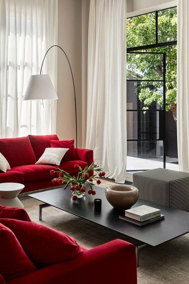 A refined living room with vivid red sofas, a low black rectangular coffee table, soft neutral carpeting, sheer floor-to-ceiling curtains, and a sculptural arched floor lamp. Large black-framed glazed doors or windows open the room visually to a leafy courtyard, while a ribbed dark media console and understated ceramic objects add texture. The look is modern, warm, and high-contrast.