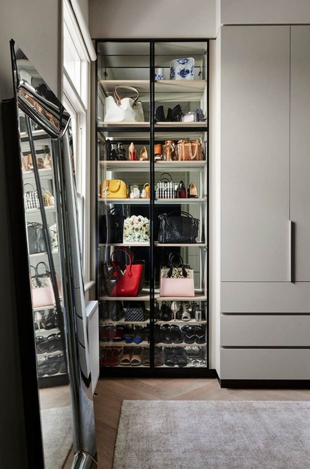 A refined walk-in wardrobe with floor-to-ceiling black-framed glass display doors, illuminated shelving filled with handbags, shoes and accessories, and adjacent matte light-grey built-in cabinetry with drawers. A large leaning mirror and neutral rug sit over warm timber flooring, creating a high-end dressing area.