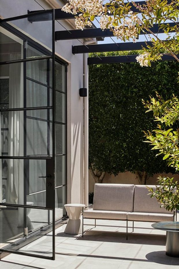 A refined indoor-outdoor living area with a white rendered wall, large black-framed glazed doors, and exposed black structural beams forming a pergola over a paved patio. A neutral upholstered sofa, sculptural side table, and low round table sit beside dense clipped greenery and flowering branches, creating a bright contemporary courtyard setting.