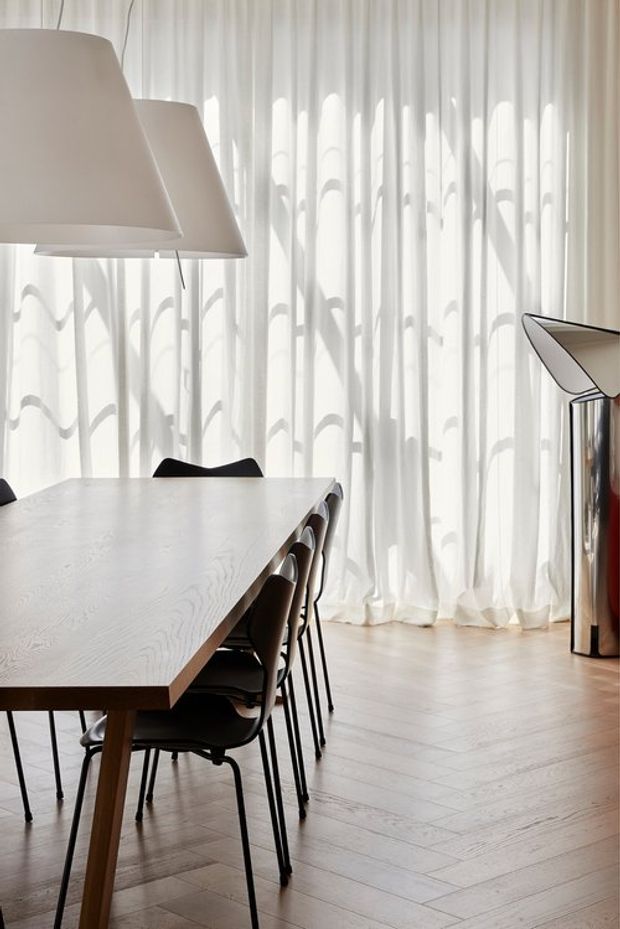 Bright, refined dining area with a long light-toned timber dining table and dark chairs arranged beneath oversized pendant lights. Full-height sheer curtains filter strong daylight across a pale herringbone timber floor, while a sculptural metallic cabinet or sideboard sits to the right.