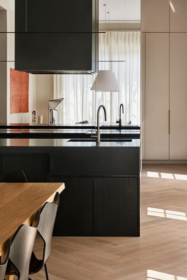 A refined modern kitchen and dining area with a dark island featuring integrated cabinetry, a black stone-look benchtop, undermount sink and twin black tapware. A large black rangehood dominates the upper frame, while reflective surfaces show pendant lighting, sheer curtains and artwork. The space includes a warm timber dining table with contemporary chairs, pale timber flooring in a herringbone pattern, and tall light-coloured cabinetry illuminated by strong natural light.