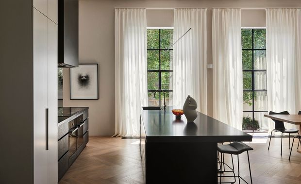 A refined open-plan kitchen and dining area with a large matte-black island, integrated dark cabinetry, and a black-framed rangehood. Tall black-framed windows with sheer white curtains bring in abundant natural light and overlook greenery. The room features warm herringbone timber flooring, neutral walls, minimalist furnishings, and sculptural decorative objects.
