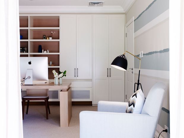 Bright, compact home office with a built-in wall of white cabinetry and open shelving, a pale wood desk with an iMac, and a light upholstered armchair beside a black-and-brass floor lamp. The room features soft grey-and-white horizontal wall stripes, white decorative ceiling mouldings, and a calm, refined palette.