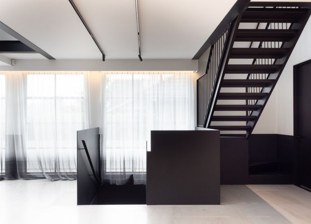 Bright contemporary interior showing a compact stairway with open black metal treads and balustrade, rising beside dark built-in cabinetry or a stair enclosure. Large floor-to-ceiling sheer-curtained windows provide abundant daylight. The space has a minimalist black-and-white palette, pale flooring, and exposed linear ceiling beams or tracks.