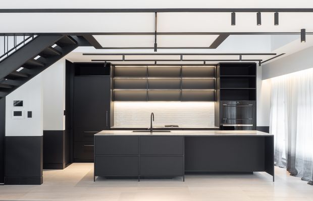 Contemporary open-plan kitchen beneath a black steel staircase, featuring a large dark island with integrated cabinetry, matching full-height joinery, a pale textured backsplash, under-cabinet lighting, and linear track and pendant lighting. Sheer curtains and bright glazing at right add natural light.