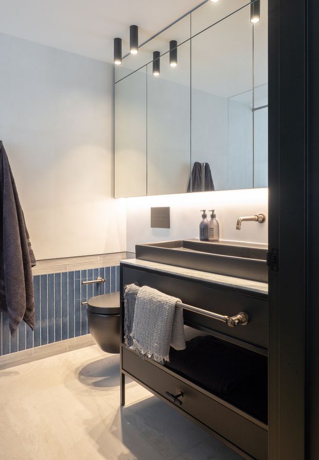A compact contemporary bathroom with a dark floating vanity, integrated basin, wall-mounted brushed metal tapware, and a large mirrored cabinet. Vertical blue wall tiles surround the toilet area, while pale stone-look flooring and warm LED lighting create a refined contrast. Matte black cylindrical pendant lights hang in front of the mirror, and towels add a practical touch.