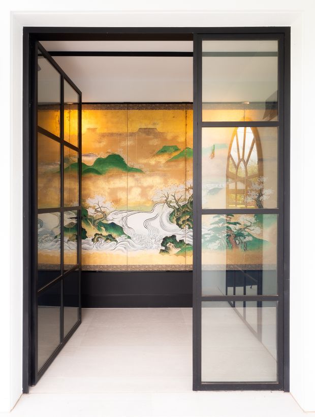 A refined interior entry or hallway framed by black metal-and-glass doors. The space features pale flooring and walls, a dark lower wall panel, and a large vibrant Japanese-inspired mural depicting stylised mountains, flowing water, and trees. Reflections in the glazing reveal additional architectural framing and a tall arched window or opening beyond.