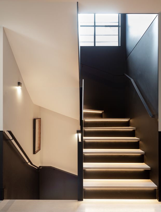 Interior view of a contemporary stairway with dark walls, floating timber-toned treads, integrated warm LED strip lighting beneath each step, black metal handrails, and a high frosted or translucent window at the upper landing. A lower stair flight, framed artwork, and wall sconces are visible to the left.