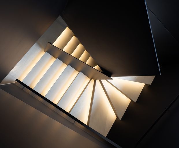 A dramatic upward view of a sculptural stairway or staircase void, with angular flights, dark structural edges, and warm concealed lighting illuminating pale geometric wall and stair surfaces. The composition emphasizes strong diagonal lines, shadow, and architectural form in a dark, high-contrast interior.