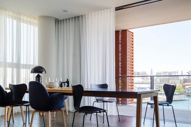 Bright contemporary dining area in a high-rise apartment, featuring a long dark timber dining table, sculptural upholstered dining chairs, a curved black floor lamp, sheer white curtains, and floor-to-ceiling glazing overlooking a waterfront urban skyline. The space has light timber flooring and a clean, airy open-plan feel.