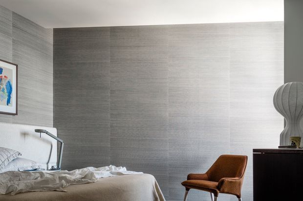 A contemporary bedroom with a low-profile bed, rumpled white bedding, a slim metal bedside lamp, a cognac leather lounge chair, and a dark timber dresser. Large grey textured wall panels create a calm, monochromatic backdrop, complemented by minimalist artwork and sculptural lighting.