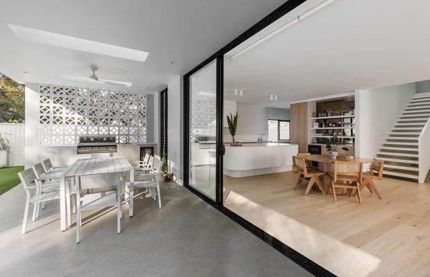Bright contemporary open-plan home with a covered outdoor dining area opening through large black-framed glazed doors to an airy interior. The interior includes a light timber dining table and chairs, a white kitchen with curved island, open shelving, pale timber flooring, and a floating staircase. A decorative breeze-block screen separates the outdoor dining area from the garden.