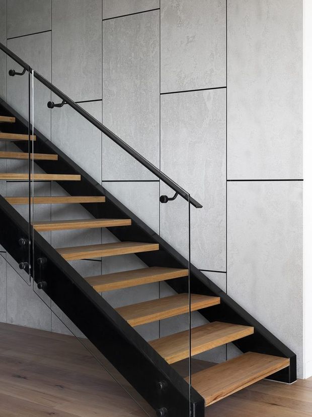 Interior view of a modern open-riser staircase with warm timber treads, black steel stringers and handrails, and clear glass balustrades. The staircase is set against a tall grey textured panelled wall, with dark timber flooring below. The composition highlights the stair structure and material contrast.