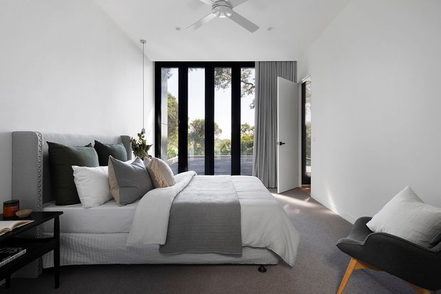 A bright contemporary bedroom with a large upholstered bed dressed in layered white and grey bedding, a dark accent headboard, grey carpet, and a black lounge chair. Full-height black-framed glazing opens toward greenery, with sheer curtains, a ceiling fan, and a partially open white door. The room has a calm, refined, neutral palette and abundant natural light.