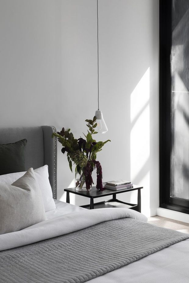 A softly lit bedroom corner featuring a grey upholstered bed with nailhead trim, layered white and grey bedding, and a small black side table holding books and a vase of foliage. A simple pendant light hangs against a white wall beside a large dark-framed window, creating strong geometric sunlight and shadow.