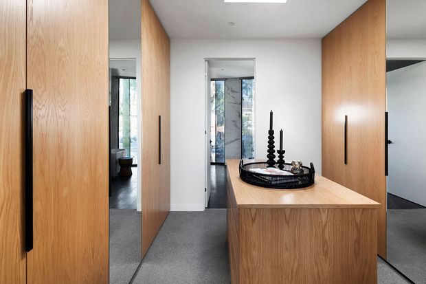 Contemporary hallway or entry area with extensive light timber veneer built-in cabinetry, tall black handles, a central timber console, and a large mirror reflecting adjacent spaces. Grey carpet, white walls and ceiling, and a simple decorative tray with black candlesticks create a clean, refined interior. A bathroom or wet area is visible through the doorway at the rear.