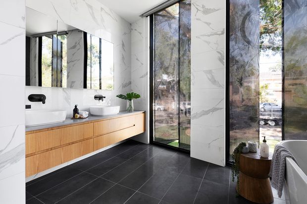 Contemporary bathroom featuring a long floating timber vanity with twin basins and wall-mounted black fixtures, a white vessel basin, full-height white marble-look wall tiles with grey veining, and large black-framed glazed openings. The room has a dark tiled floor, a freestanding white bathtub at the right, and abundant natural light with views to trees and the street outside.