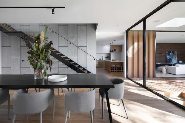A bright, open-plan contemporary dining and living area with a large black dining table and upholstered chairs in the foreground. A sculptural floral arrangement sits on the table. Behind it, a black metal stair with open treads runs along a large grey tiled feature wall, while a compact kitchen and wine storage are visible beyond. Black-framed glass partitions separate the dining area from a sunlit living room with timber flooring and vertical timber screening. A skylight and recessed lighting add to the airy, refined interior.