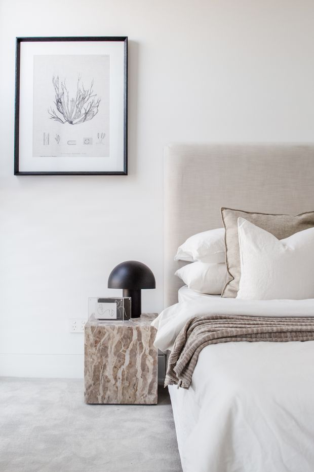 A softly styled bedroom corner with a white upholstered bed, layered white and taupe pillows, and a textured taupe throw. A sculptural dark table lamp and small decorative objects sit on a chunky natural stone bedside table. The room has pale walls, light grey carpet, and restrained framed botanical artwork, creating a calm, refined neutral scheme.