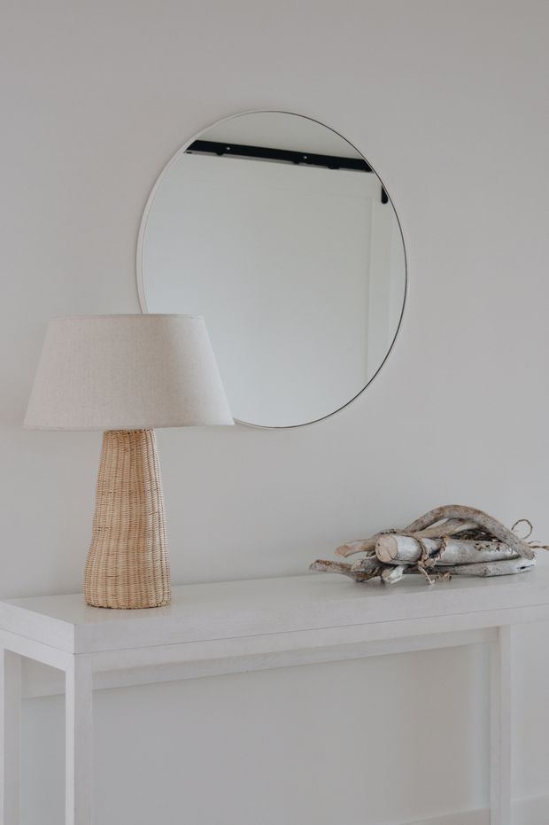 A softly styled minimalist interior vignette featuring a white console table against a light grey-white wall, a round wall mirror, a woven natural-fibre table lamp with a white fabric shade, and a small sculptural driftwood arrangement. The palette is pale, neutral, and understated.
