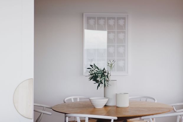 A bright, minimalist dining area with a round light-wood dining table surrounded by white chairs. The space has pale walls, a simple framed botanical artwork, and a small vase with greenery as the central table arrangement.