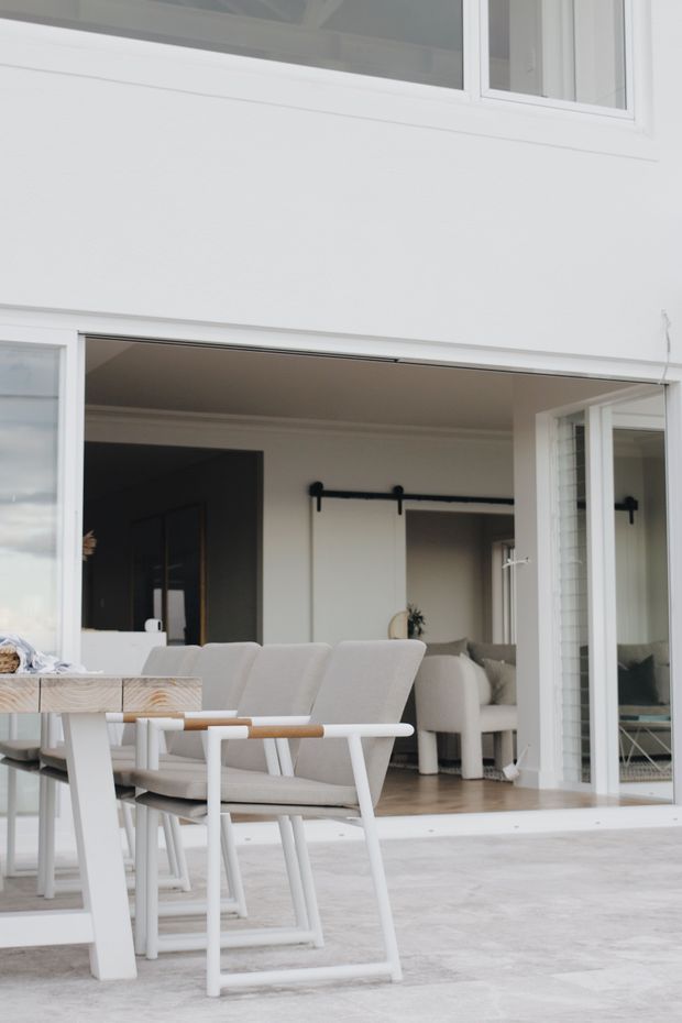 A bright, minimalist residential dining and living area viewed from a pale outdoor terrace through large open glazed doors. The foreground has a light timber dining table with white-painted legs and upholstered light-grey dining chairs. Inside, a neutral sofa, light timber flooring, white walls and ceilings, and a black barn-style sliding door track create a clean contemporary interior. The composition also shows white-framed glazing and a calm, muted neutral palette.