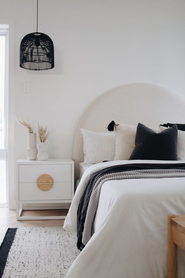 Bright, softly styled bedroom featuring a large rounded upholstered bedhead, layered white and charcoal bedding, a white bedside table with timber drawer pull, neutral rug, and a black woven pendant light. The palette is calm and predominantly white, cream, grey, and black with natural-textured accents.