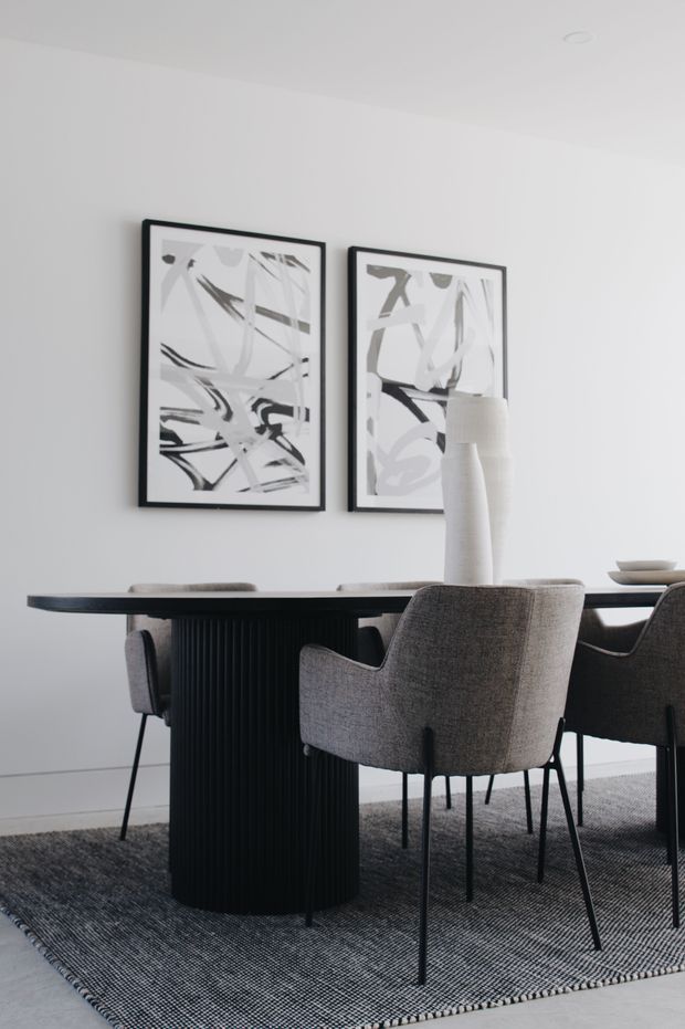 A refined dining room with a long dark dining table, upholstered grey dining chairs, a tall white sculptural vase, monochrome abstract artwork, and a large textured grey rug. The space has white walls, a clean contemporary composition, and a restrained neutral palette.