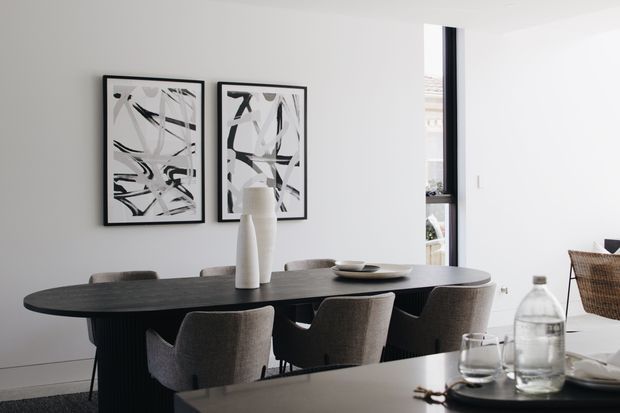 Contemporary dining area with a long dark dining table surrounded by upholstered chairs, set against white walls with two monochrome abstract artworks. A tall vase and shallow dish form a simple centrepiece, while a large window brings in daylight. Part of a dark kitchen or sideboard surface is visible in the foreground.