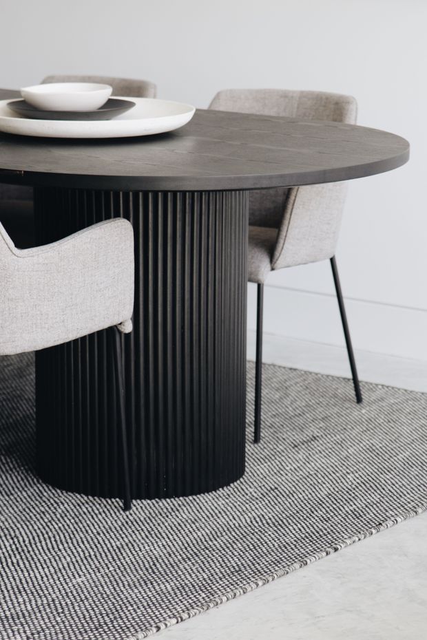 Close-up interior product-style photograph of a round dining table with a dark grey stone-look top and vertically fluted black pedestal base, surrounded by light grey upholstered dining chairs. White ceramic plates and bowls sit on the tabletop. The setting is a restrained contemporary dining area with a monochrome palette, light wall, textured grey rug, and pale hard flooring.