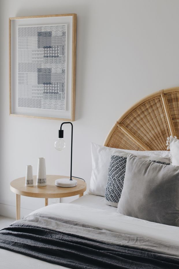 A softly styled bedroom vignette with a light neutral wall, framed monochrome artwork, a round light-wood bedside table, minimalist black lamp, ceramic vessels, and a bed dressed in white, grey, and charcoal textiles. A large woven rattan fan-shaped headboard adds a natural textural feature.