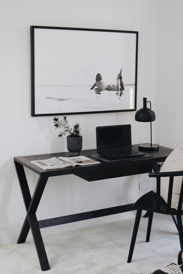 A minimalist home workspace with a black trestle-style desk, open laptop, desk lamp, potted plant, and open magazine. The desk sits against a white wall with a large monochrome framed photograph above it; a black chair and pale carpet complete the scene.