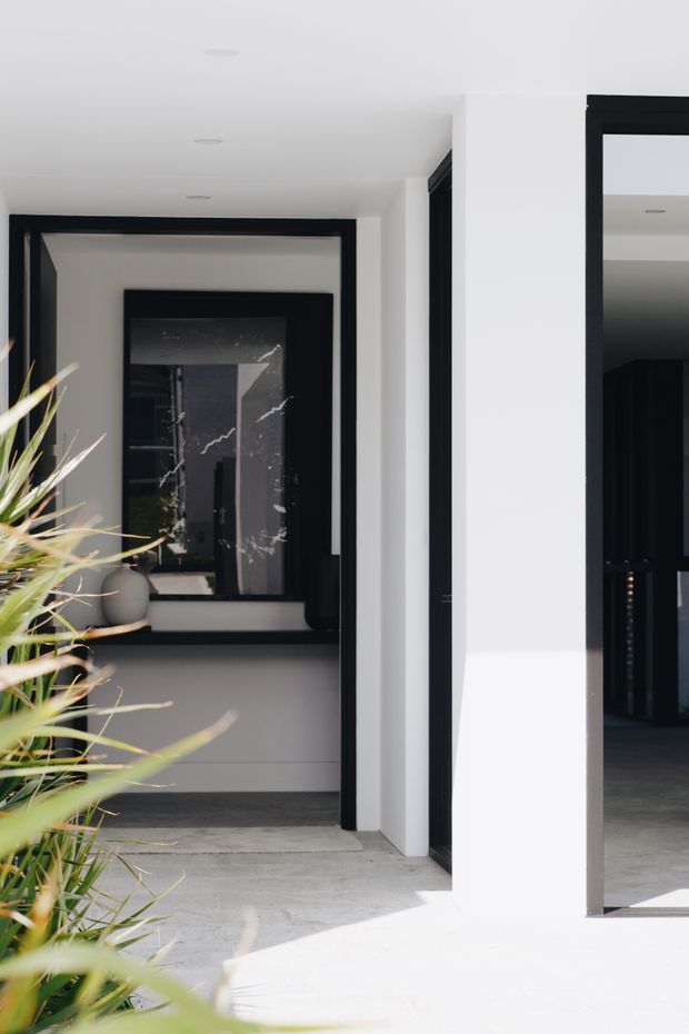 Bright minimalist entryway with white walls, pale concrete flooring, and strong black-framed internal doorways and glazing. A large mirror or reflective dark glass panel extends the view into the adjoining spaces, while a potted architectural plant softens the monochrome scheme. The composition emphasizes crisp geometric lines, natural light, and a refined contemporary interior.