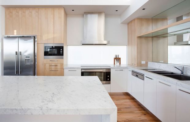 Bright contemporary kitchen with a large white stone island, pale timber cabinetry, white lower cabinets, stainless-steel appliances, mirrored splashback, and warm timber flooring. The space has clean lines, integrated storage, and abundant reflective surfaces.