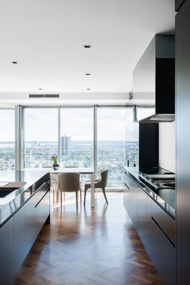 A bright, contemporary kitchen with dark full-height cabinetry and integrated appliances framing a dining area. Floor-to-ceiling glazing provides expansive elevated city views, while a warm herringbone timber floor contrasts with the minimalist white ceiling and pale walls.