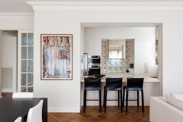 A bright, refined open-plan interior showing a dining area in the foreground and a compact kitchen beyond. The kitchen features white cabinetry, a pale benchtop, stainless-steel appliances, a patterned tiled splashback, and a narrow breakfast bar with three black bar stools. White walls with decorative crown moulding, glazed internal doors, framed artwork, and herringbone timber flooring contribute to a classic yet contemporary character.
