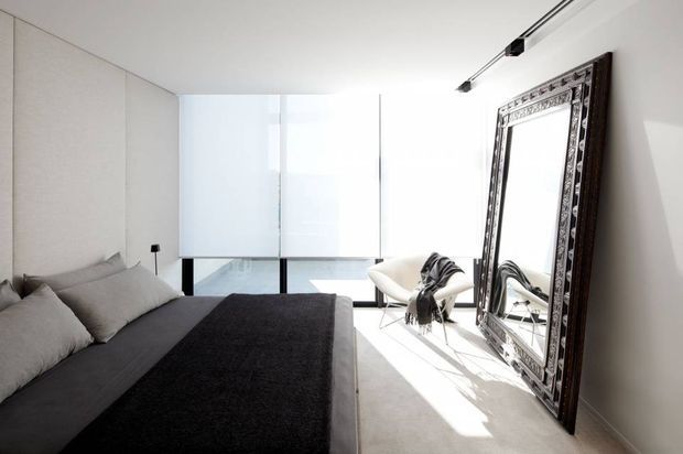 Bright contemporary bedroom with a low-profile bed dressed in charcoal and grey textiles, pale upholstered or textured wall panels, and floor-to-ceiling glazing at the far end. Two sculptural lounge chairs sit beside a large ornate dark-framed leaning mirror, while a minimalist track light runs along the ceiling. The room has a clean monochromatic palette and strong natural light.